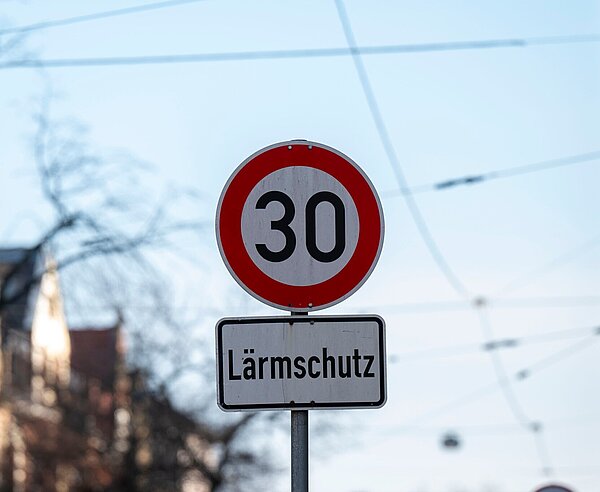 Aufnahme 30er Zone- und "Lärmschutz"-Schild vor Himmel, Bäumen und Oberleitungend der Bahn.