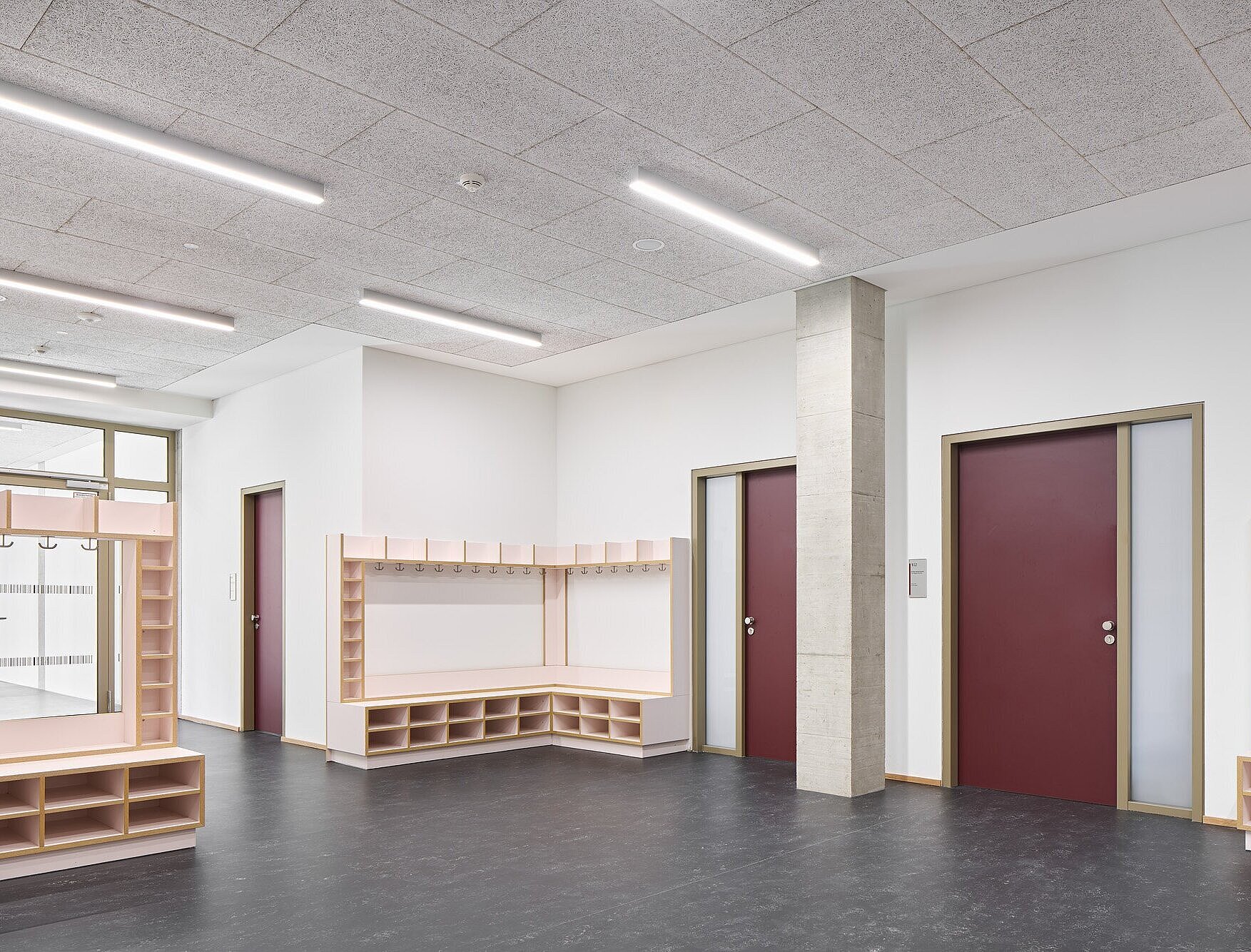Zu sehen ist nochmals einer der Lernflure mit direktem Blick auf die roten Türblätter der Klassenräume. Daneben befinden sich die Garderoben als Einbaumöbel aus Holz mit rosafarbenen Flächen. Der Flur ist mit einem dunkelgrauen Linoleumboden sowie einer hellen Heraklithdecke ausgestattet