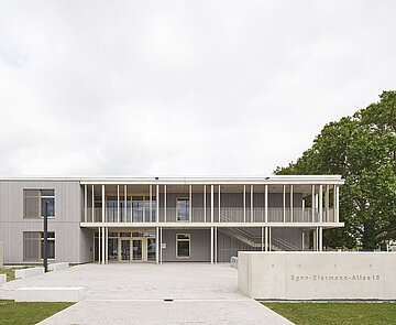 Das Bild zeigt die Südwestfassade mit heller Holzverkleidung, Balkon und dem Eingangsbereich mit Schriftzug.