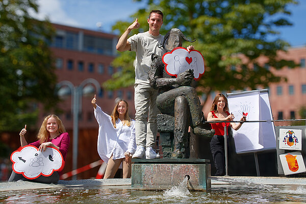 Das Bild steht symbolisch für die Fortbildung für Ehrenamtliche. Zu sehen sind vier Personen vor dem Narrenbrunnen in Karlsruhe, die Plakate mit Motiven rund um ehrenamtliches Engagement hochhalten und ein „Daumen hoch" geben.