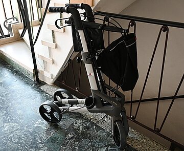 Ein Rollator steht zusammengeklappt an einem Treppengeländer im Hausflur.