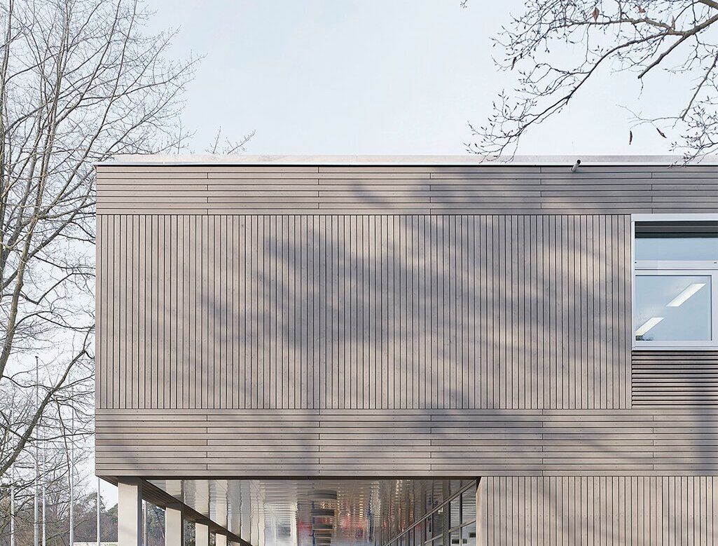 Das Bild zeigt eine Detailansicht der grau lasierten Holzfassade und ein überdachter Weg für die Schüler, der durch das darüberliegende, vorspringende Geschoss entsteht.