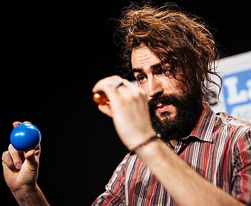 Ein FameLab-Teilnehmer hält eine blaue und eine rote Kugel hoch