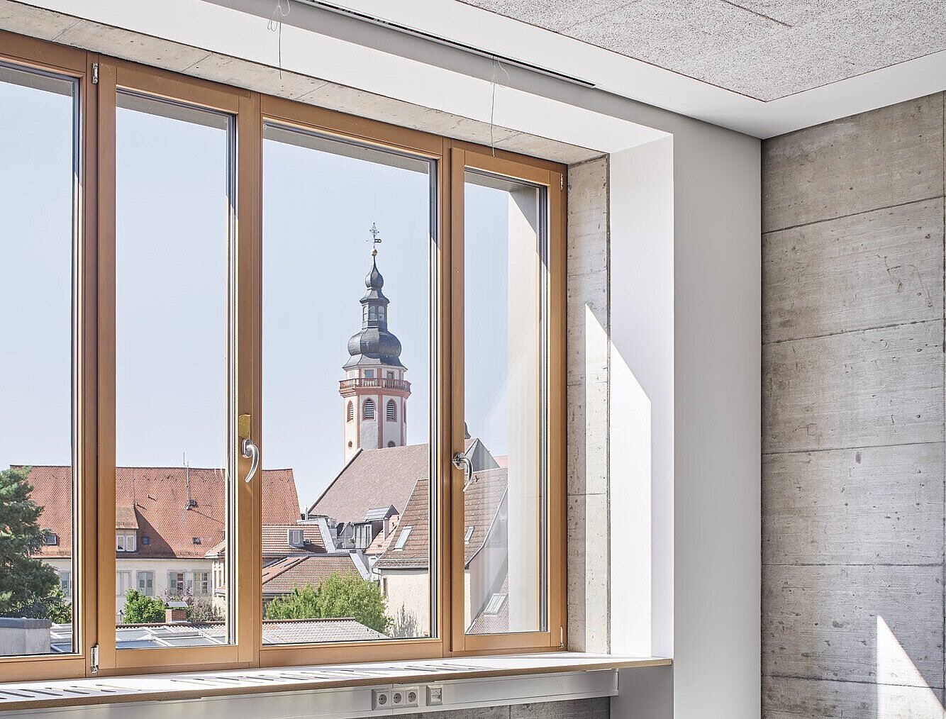 Das Bild zeigt ein Detail der Fenster mit Holzrahmen und Ausblick auf den Kirchturm. Unterhalb der Fenster ist eine dezente, weiße Heizung angeordnet. Der graue Linoleumboden mit Holzleisten und die helle Heraklithdecke ergänzen das Raumdetail.