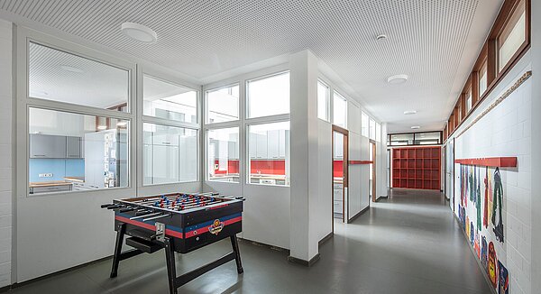 Zu sehen ist der helle, moderne Flurbereich mit Kautschukboden und abgehängter Gipskarton-Lochdecke, der teilweise als Spielbereich mit Tischkicker genutzt wird.