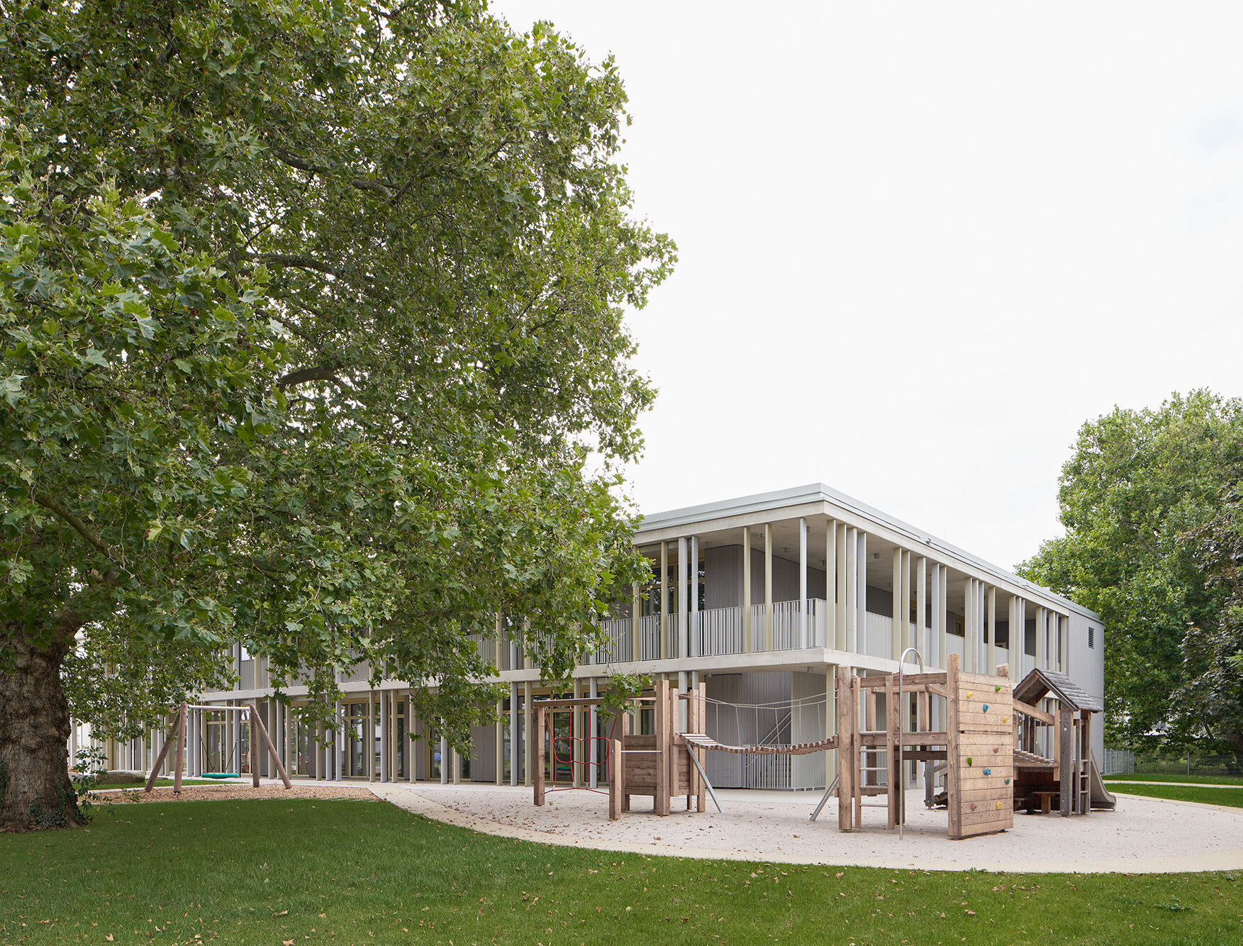 Zu sehen ist der Spielgarten und die Ostansicht des Gebäudes, die teilweise von einem großen Baum verdeckt wird. 