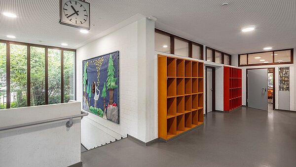 Das Bild zeigt den hellen Flurbereich mit erneuerten Türblättern der Gruppenräume, grauem Kautschukboden und weißer Akustikdecke. Die Einbauschränke und ein Bild am Treppenabgang setzen farbliche Akzente.