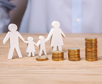 vier unterschiedlich hoche Stapel Kleingeld, dahinter eine Silhouette einer Familie aus dicker weißer Pappe