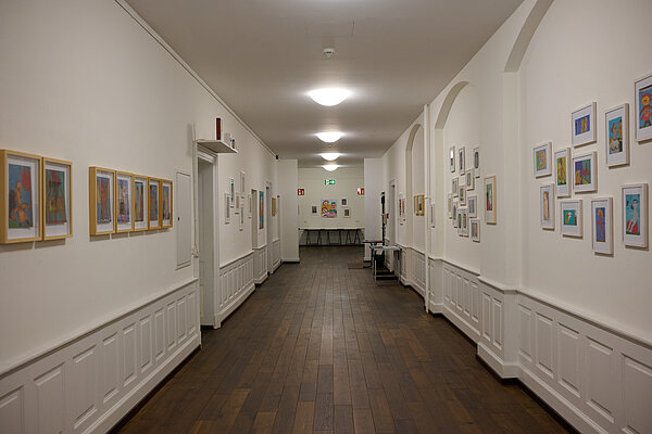 Blick in einen weißen, länglichen Ausstellungsraum mit Bildern an den Wänden