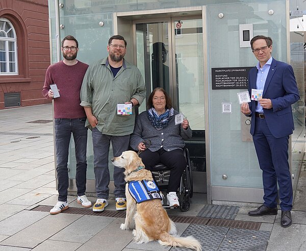 Am Karlsruher Marktplatz testen (v.l.n.r.) Christian Höglmeier (technischer Geschäftsführer der VBK), Ulrike Wernert (kommunale Behindertenbeauftragte der Stadt Karlsruhe), Artur Bundnik (Projektstelle Web-App „Karlsruhe barrierefrei“) und Marcus Albert (System-Administrator „Karlsruhe barrierefrei“) die neuen QR-Codes an den Stadtbahntunnel-Aufzügen, über die die Nutzer*innen schnell und direkt auf die Seite „Karlsruhe barrierefrei“ gelangen.