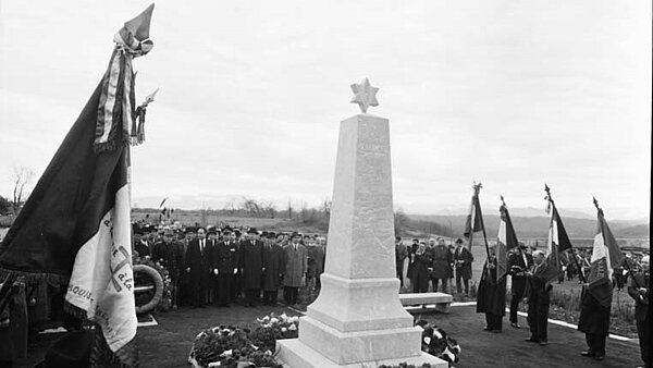 Ansicht des mit Kränzen geschmückten Mahnmals. Um das Mahnmal Fahnenträger und die Menge der Teilnehmenden stehend.