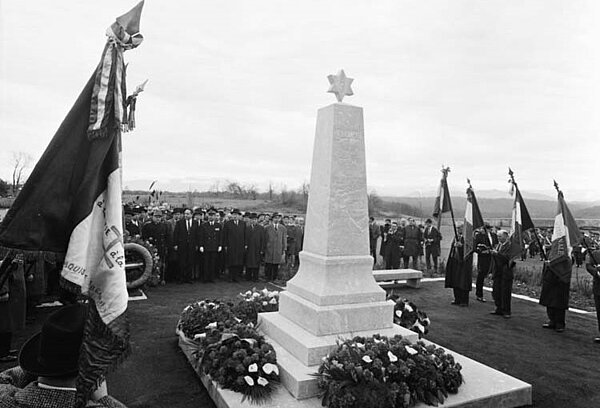Ansicht des mit Kränzen geschmückten Mahnmals. Um das Mahnmal Fahnenträger und die Menge der Teilnehmenden stehend.
