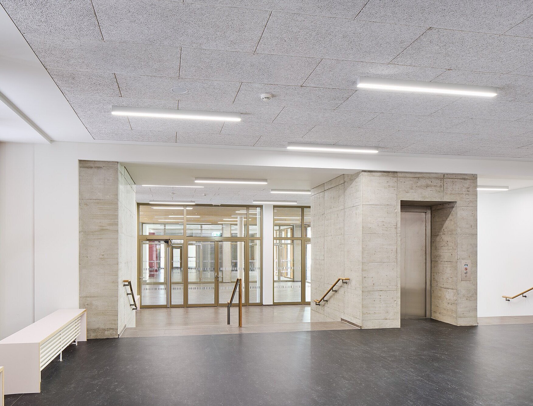 Das Bild zeigt die Bestandsaula mit einem zentralen Personenaufzug. Auf beiden Seiten des Aufzugs befinden sich kleine Treppenflächen. Die Decke ist mit weißem Heraklithplatten verkleidet. Die Aula grenzt an die innenliegende Pausenfläche an. 