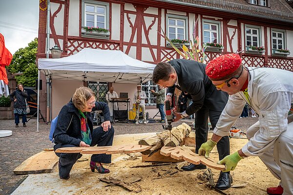 Der Holzbildhauer und Aktionskünstler Guntram Prochaska gestaltet auf dem Rathausplatz gemeinsam mit der bisherigen Grötzinger Ortsvorsteherin Karen Eßrich und ihrem Nachfolger Jens Jägle ein Kunstwerk. 