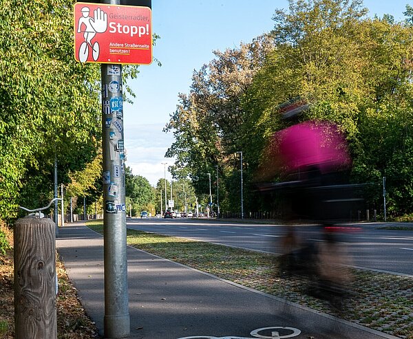  Blick auf den Fahrradstreifen am Adenauerring mit Hinweisschild auf Geisterradler und Fahrradfahrer der entgegen der zulässigen Fahrtrichtung auf dem Radfahrstreifen unterwegs ist.