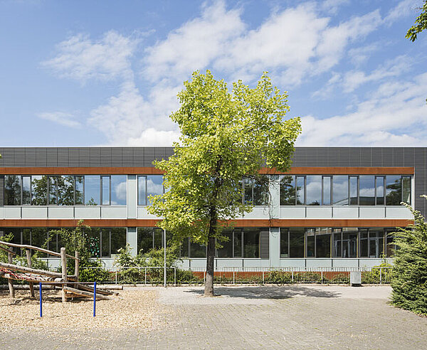 Das Bild zeigt den Schulhof und die anthrazitfarbene Südfassade, die mittig von einem kleinen Baum bedeckt ist.