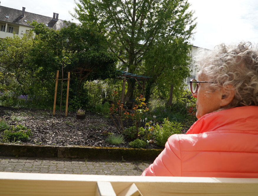 Frau von hinten auf Bank sitzend im Vordergrund. Garten im Hintergrund.