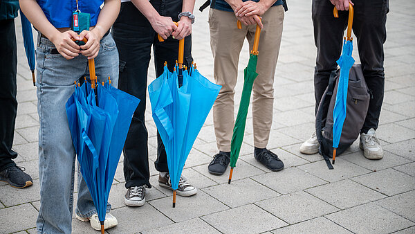 Das Bild zeigt die Beine von vier Personen, die Schirme in der Hand halten.  Diese Schirme symbolisieren im Rahmen einer spontan Aktion, so genannter FreshMob, die Entsiegelung der Innenstadt. 