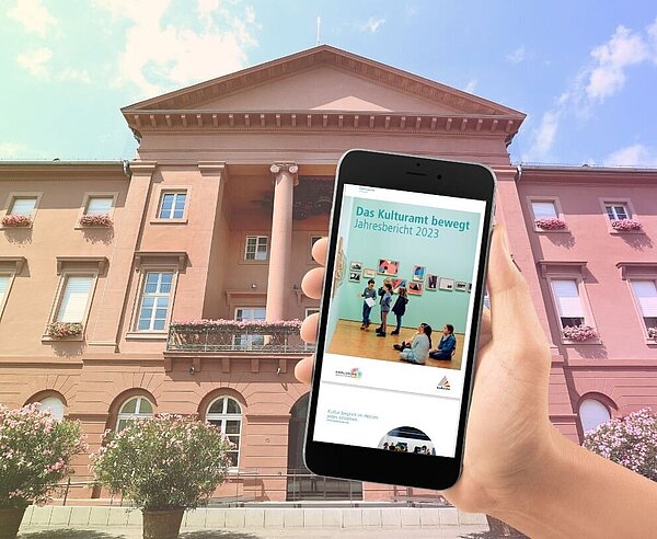 Eine Hand hält ein Smartphone, auf dem der Kulturamt Jahresbericht 2023 geöffnet ist vor das Rathaus Karlsruhe. Das Bild ist eine Foto-Collage und das Rathaus im Hintergrund wurde mit einem warmen Farbverlauf von gelb in rot leicht verändert