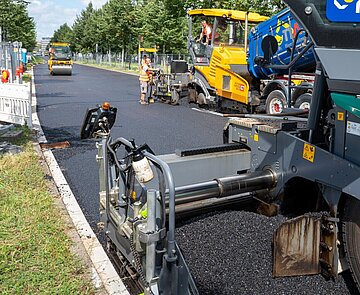 Bei der Baustelle an der Ludwig-Erhard-Allee wird zur Fahrbahnsanierung ein neuartiger Niedertemperaturasphalt auf der Fahrbahn aufgebracht.