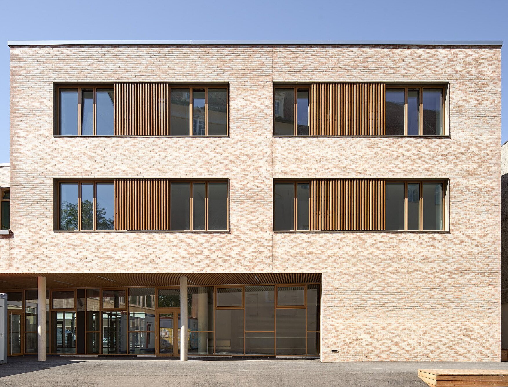 Zu sehen ist die Ostfassade mit beigefarbenem Klinkermauerwerk, das sich an die Farbgebung der umliegenden Gebäude anlehnt. Die Fenster bestehen aus Metallelementen mit Holzlamellen.