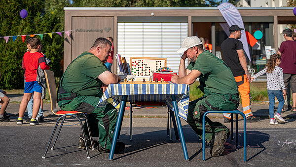 Zwei Männer sitzen an einem Tisch und spielen Schach. Im Hintergrund sind Kinder und Erwachsene zu sehen.