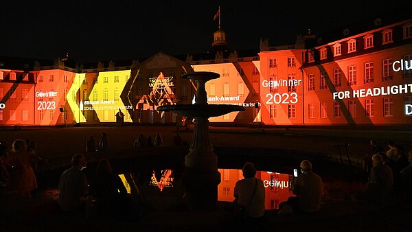Auf dem Schloss wird das Stück "For headlights crave hindsight too" des Kluster Kollektiv angekündigt. Das Wasser eines Brunnen reflektiert die Farben.