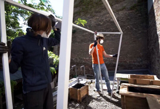 Zwei Personen halten Gerüst aus Holzlatten im Hinterhof. Hochbeete stehen auf dem Boden