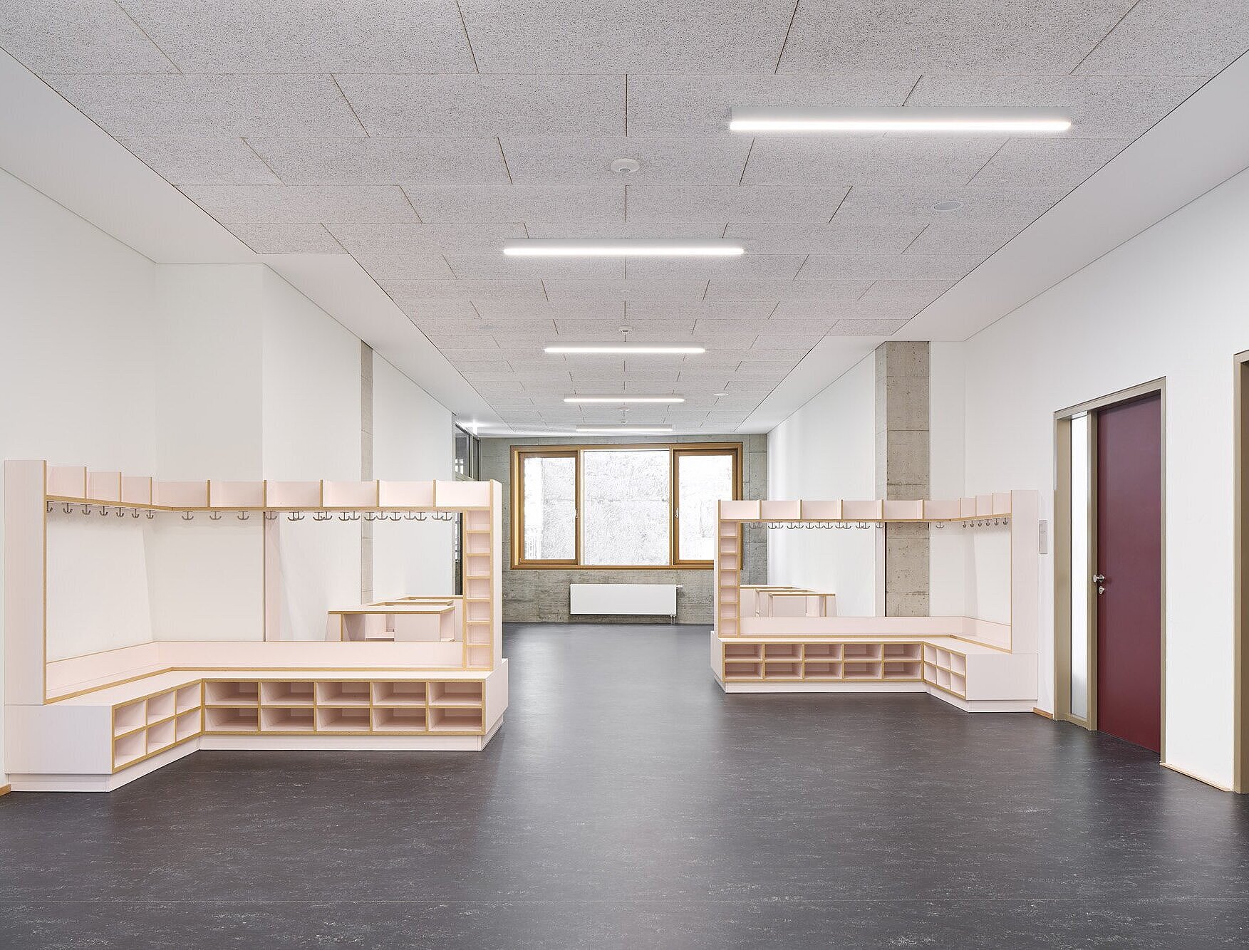 Zu sehen ist einer der Lernflure mit dunkelgrauem Linoleumboden und heller Heraklithdecke. Auffällig sind die hölzernen Garderoben als Einbaumöbel mit rosafarbenen Flächen sowie die roten Türblätter der Klassenräume.