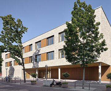 Die Abbildung zeigt die west-südliche Fassade des Gebäudes mit beige­farbenem Klinkermauerwerk, einem Flachdach und Metallfenstern mit Holzlamellen. Die Gestaltung ist farblich an die umliegende Bebauung angepasst