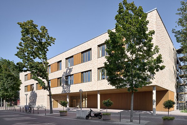 Die Abbildung zeigt die west-südliche Fassade des Gebäudes mit beige­farbenem Klinkermauerwerk, einem Flachdach und Metallfenstern mit Holzlamellen. Die Gestaltung ist farblich an die umliegende Bebauung angepasst