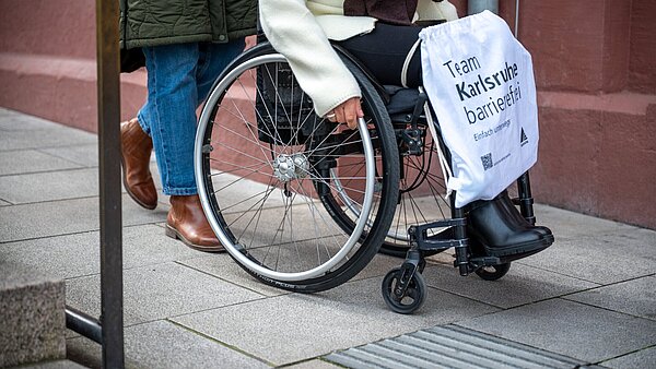 Seitenansicht einer Begleitperson, die eine Person im Rollstuhl sitzend schiebt. Diese hält einen Stoffbeutel mit der Aufschrift "Team Karlsruhe barrierefrei"