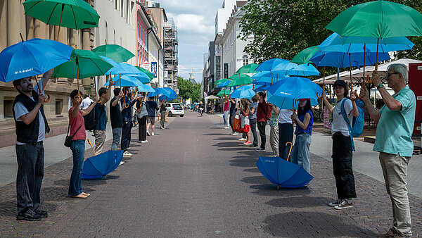 Das Bild zeigt die Herrenstraße. Links und rechts ist eine Allee von Menschen zu sehen, die blaue und grüne Schirme in die Luft halten. Diese Schirme symbolisieren schattenspende Bäume, die im Rahmen einer spontanen Aktion, so genannter FreshMob die Notwendigkeit verdeutlichen sollen, dass im Zuge des Klimawandels mehr Bäume in der Innenstadt benötigt werden.