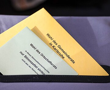 Stimmzettelumschläge für die Wahl des Gemeinderats und die Wahl des Ortschaftsrats halb im Schlitz einer Wahlurne.