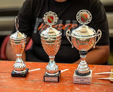 Drei Pokale stehen auf einem Tisch und werden mit Schriftzügen beklebt.