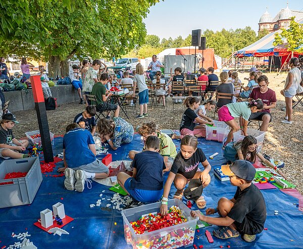 Bürgermeister Dr. Albert Käuflein wird von Kindern bei einem Besuch im Camp der Möglichkeiten interviewt. Im Vordergrund mehrere Kinder an der Station mit Legospielsteinen auf dem Boden sitzend.