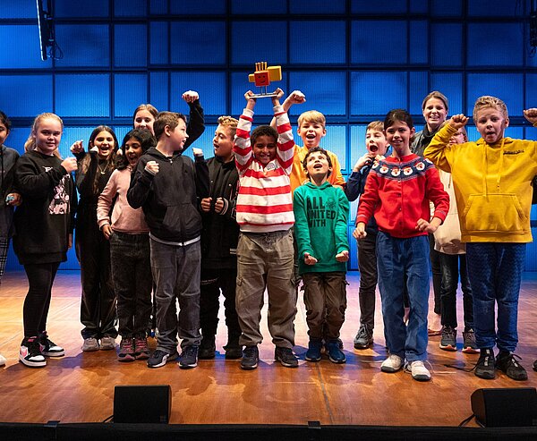 15 Kinder jubeln blickend in die Kamera. Kind in der Mitte hebt Pokal hoch.
