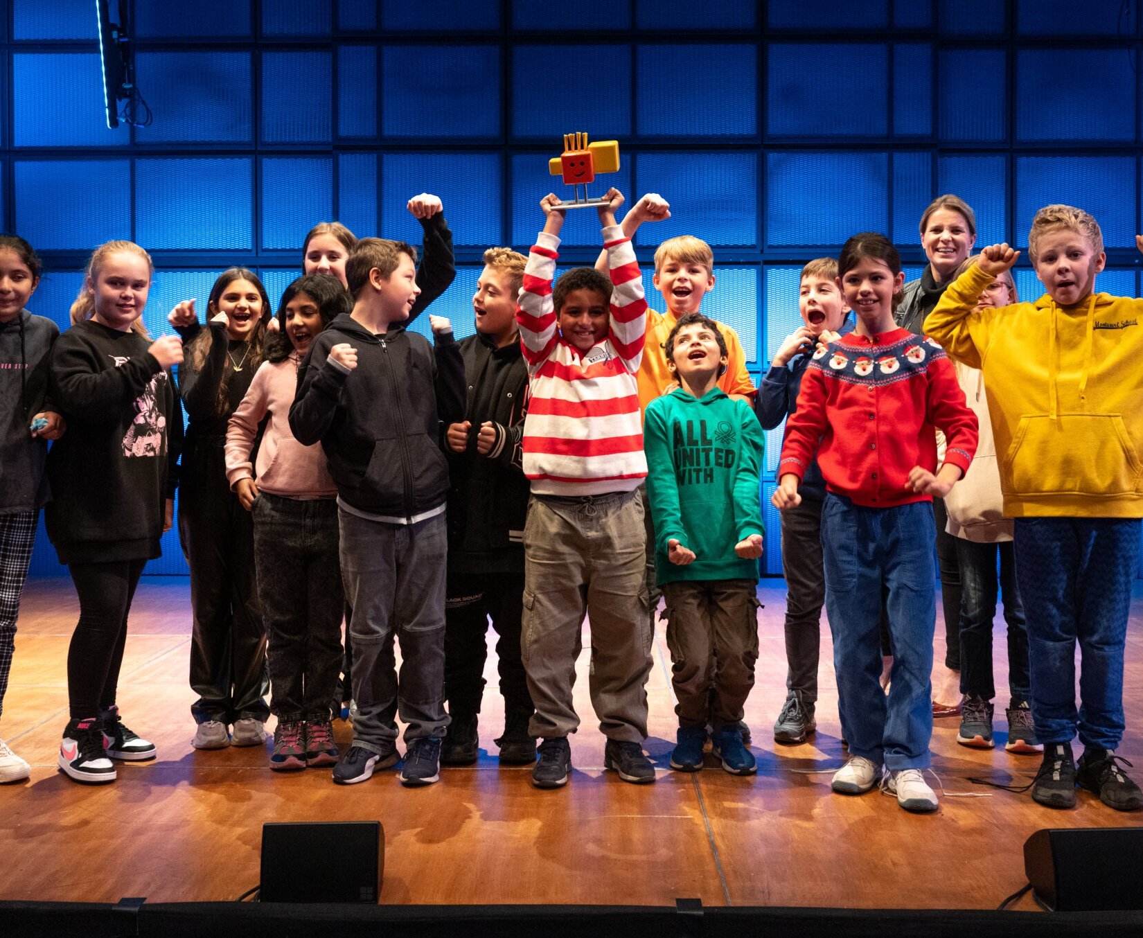 15 Kinder jubeln blickend in die Kamera. Kind in der Mitte hebt Pokal hoch.