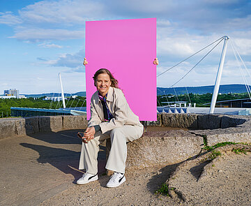 Frau in Hosenanzug sitzt auf einem Stein und im Hintergrund sieht man die Spitze der Europahalle
