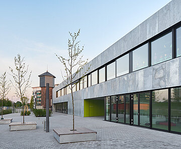 Zu sehen ist die helle Nordfassade mit großen Fensterflächen, Sitzgelegenheiten und im Hintergrund der Wasserturm.