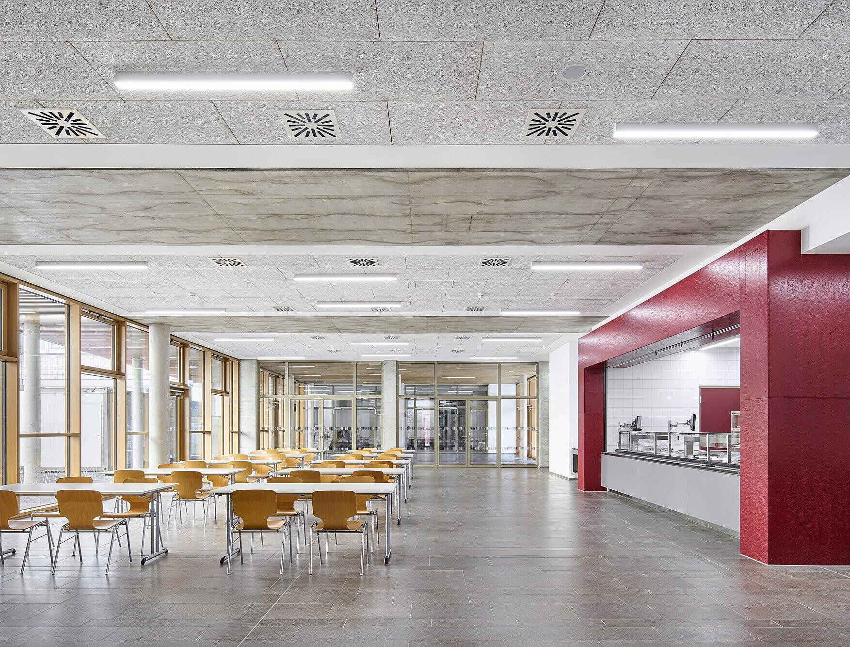 Zu sehen ist der helle Speisesaal mit großen bodentiefen Fensterflächen. Der Boden ist aus Porphyrstein gefertigt. Eine Ausgabetheke mit roten Akzenten prägt den Raum. 