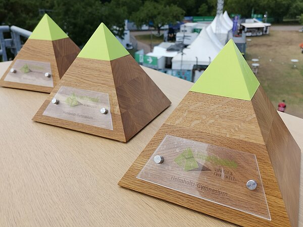 Zu sehen sind drei kleine Pyamiden aus Holz mit grüner Spitze, die Hauptpreise der Auszeichnung.