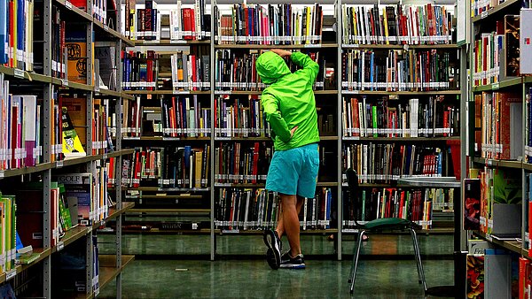 Ein Jugendlicher steht mit neongrüner Jacke und blauer kurzer Hose vor einem vollen Bücherregal. Er ist von hinten zu sehen.