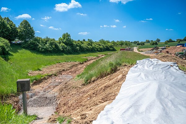 Das bestehende Rückhaltebecken (links im Bild) wird um den Bereich rechts der Plane erweitert.