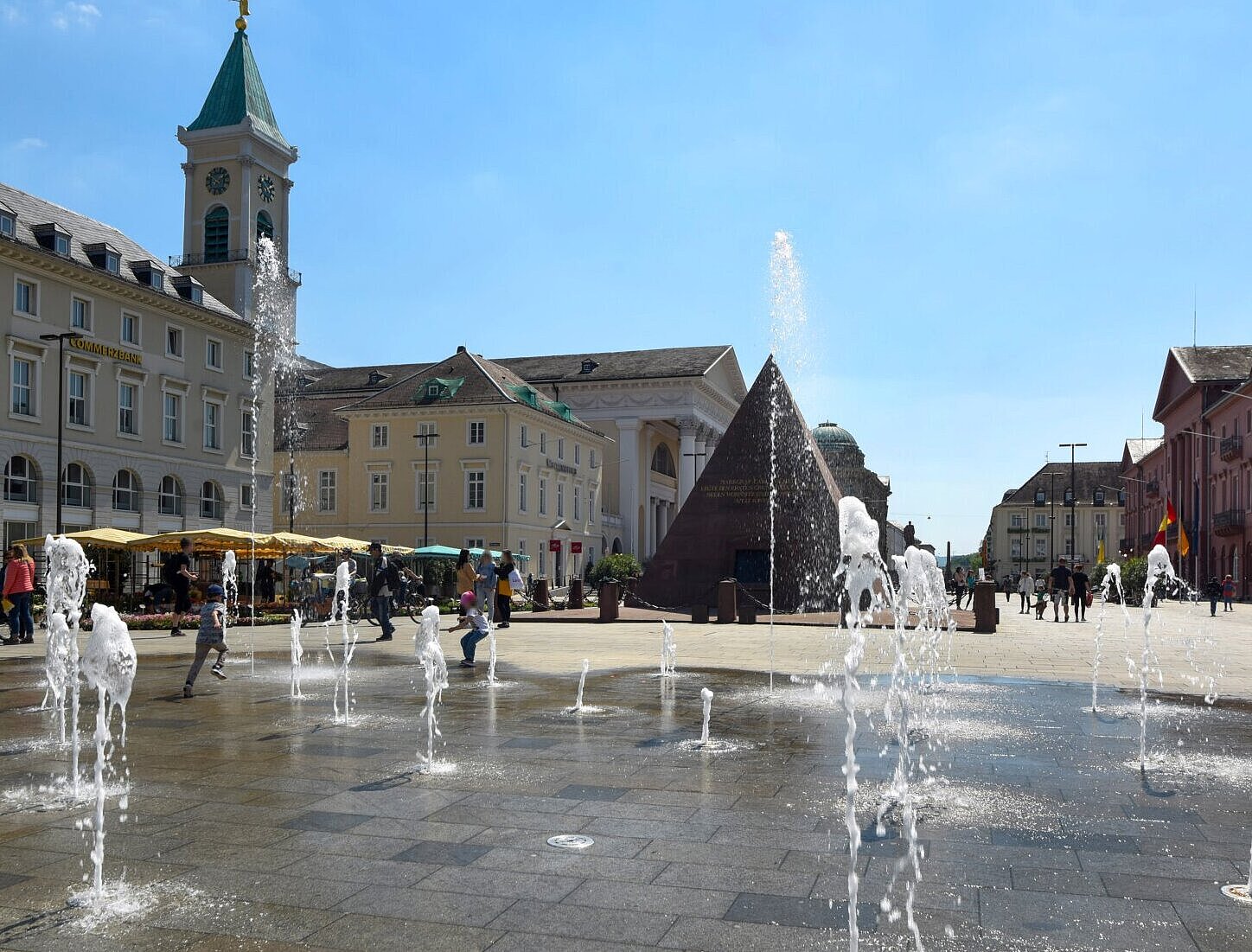 belebter Marktplatz mit Menschen und Wasserspiel im Sommer