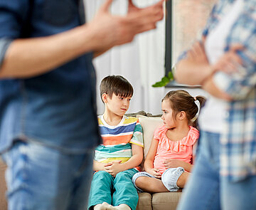Zwei Kinder die auf dem Sofa sitzen während sich die Eltern im Vordergrund streiten.