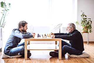 Das Bild zeigt einen jüngeren Mann, der mit einem älteren Mann an einem Tisch sitzt und Schach spielt.