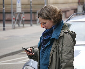 Das Bild zeigt eine Frau, die auf der Straße neben einem parkenden Auto steht, mit einem Smartphone in der Hand.