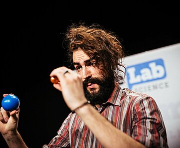 Ein FameLab-Teilnehmer hält eine blaue und eine rote Kugel hoch