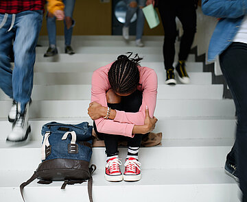 Jugendliche Person sitzt mit angezogenen Beinen und Kopf auf den Knien auf der Schultreppe, während Mitschülerinnen und Mitschüler vorbeilaufen.